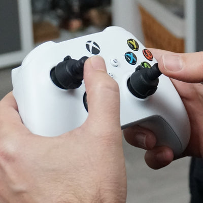 Person holding a white gaming controller with Thumb Soldiers Navigation kit attachments and a blurred indoor background