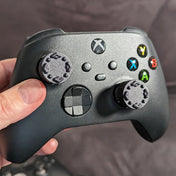 Black Xbox controller held in a hand with Thumb Soldiers' Studs thumb grips on the thumbsticks and a blurred background 