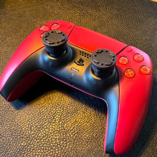 Red and black playstation gaming controller on a textured surface with Thumb Soldiers' Studs thumb grips on the thumbsticks.