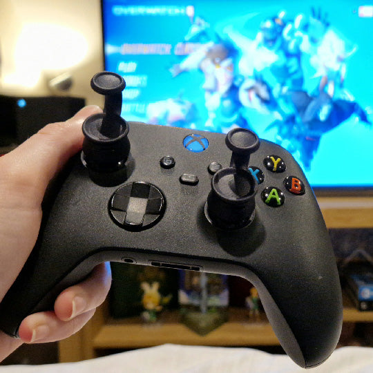 Person holding a black Xbox controller  with Thumb Soldiers' Step Up thumb grips on the thumbsticks, infront of a blurred screen with gameplay in the background.
