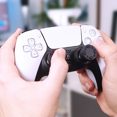 Close-up of hands holding a white gaming controller with Thumb Soldiers Studs attachments in front of a blurred indoor background