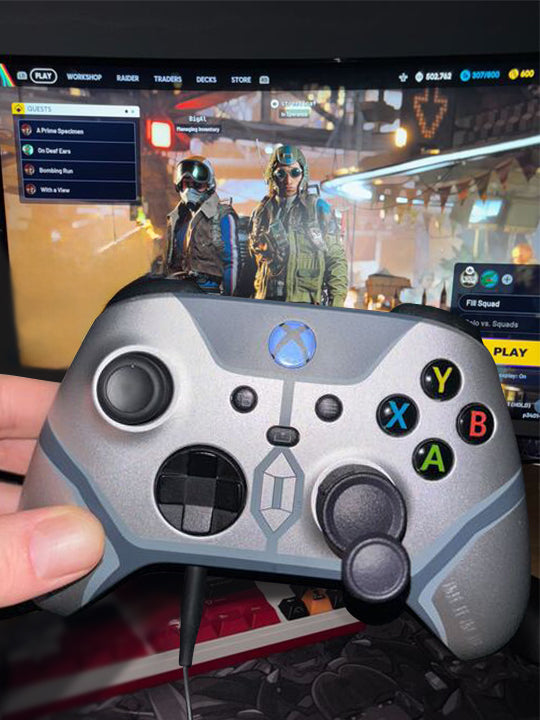 Person holding a grey Razer controller  with Thumb Soldiers' Step Up thumb grip on the thumbsticks, infront of a screen with Arc Raiders gameplay in the background.