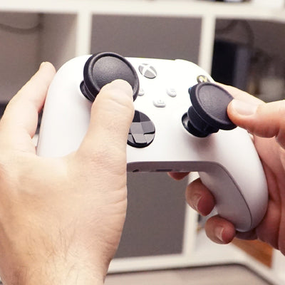 Close-up of hands holding a white gaming controller with Thumb Soldiers Shrooms kit attachments and a blurred indoor background